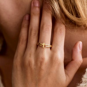 May include: A gold-toned open ring featuring a central cross design. The band splits into two parallel lines, with one end terminating in a small, clear, round-cut gemstone and the other end featuring a small, clear, round-cut gemstone.