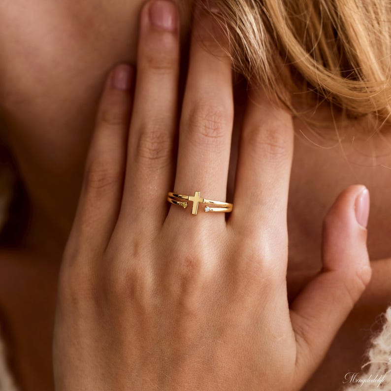 May include: A gold-toned open ring featuring a central cross design. The band splits into two parallel lines, with one end terminating in a small, clear, round-cut gemstone and the other end featuring a small, clear, round-cut gemstone.