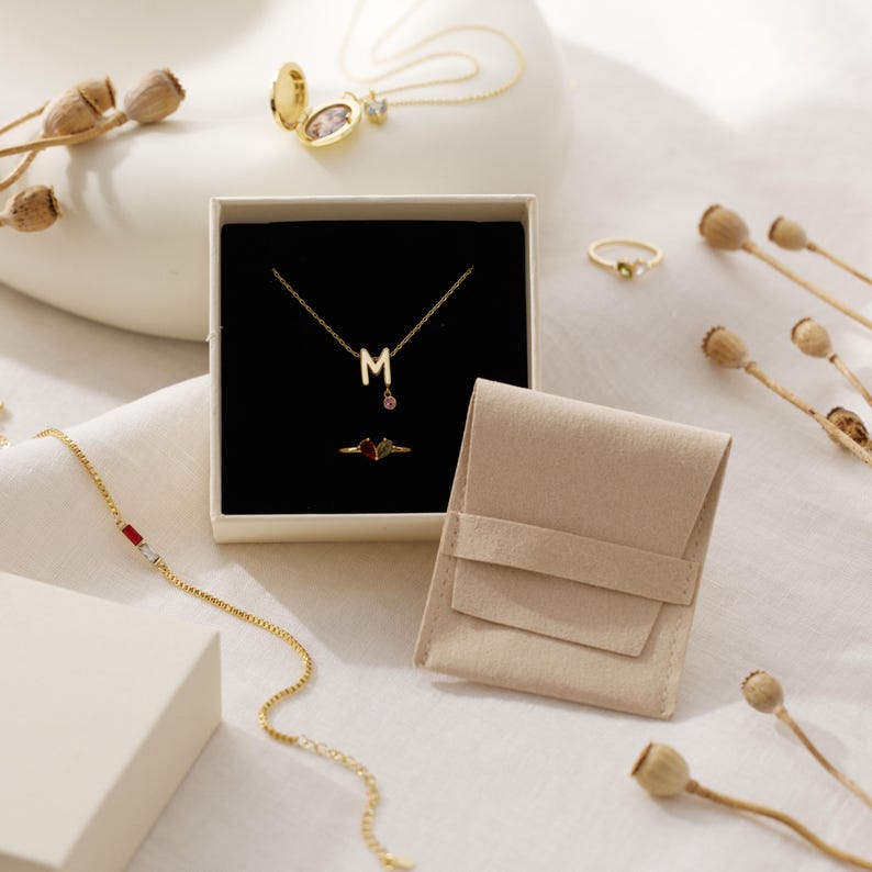 May include: A gold necklace with a letter M pendant and a small pink stone, a gold ring with two colored stones, a gold chain bracelet with red and white stones, a gold locket necklace, and a gold ring with a green stone in a beige box.