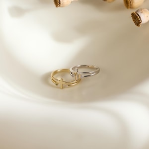 May include: Two rings, one gold-toned and one silver-toned, each featuring a delicate cross emblem at the center. The bands are thin and split into two parallel lines, with a small, clear stone set on either side of the cross on each ring.