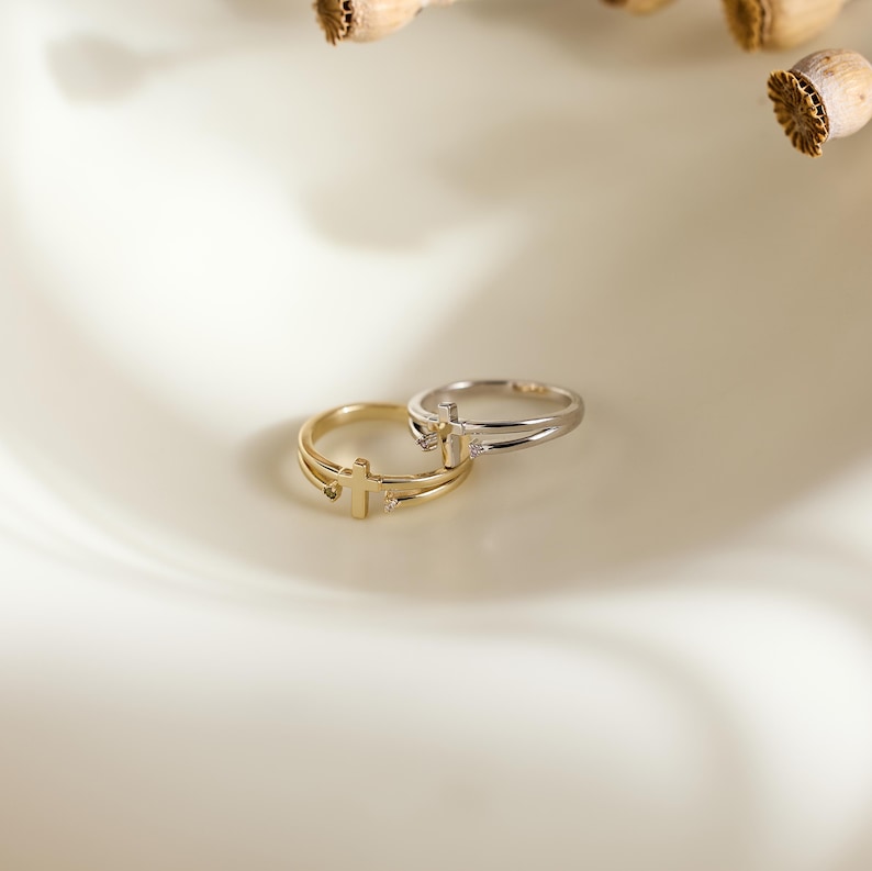 May include: Two rings, one gold-toned and one silver-toned, each featuring a delicate cross emblem at the center. The bands are thin and split into two parallel lines, with a small, clear stone set on either side of the cross on each ring.