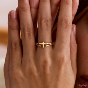 May include: A gold-toned open ring featuring a central cross design. The band is split, with one end terminating in a small, clear, round-cut stone and the other end featuring a simple, polished gold cross element at the center of the finger.