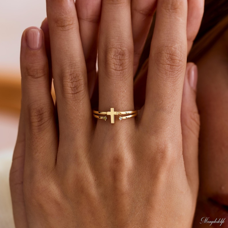 May include: A gold-toned open ring featuring a central cross design. The band is split, with one end terminating in a small, clear, round-cut stone and the other end featuring a simple, polished gold cross element at the center of the finger.