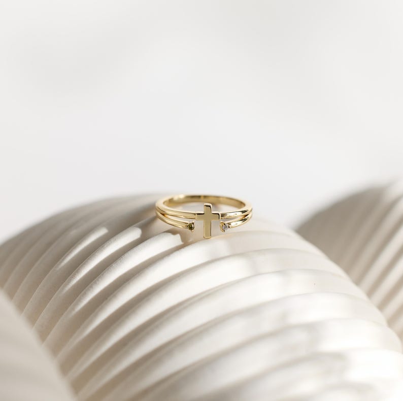 May include: A gold-toned ring featuring a double-band design that converges at the center into a small, upright cross motif. The ring is accented with two tiny, clear stones set at the points where the bands meet the cross, creating a delicate look.