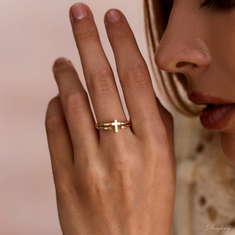 May include: A gold ring featuring a central cross design and two small clear stones set on either side of the cross. The band is thin and polished, with the cross element positioned horizontally across the finger. The metal has a warm yellow tone.
