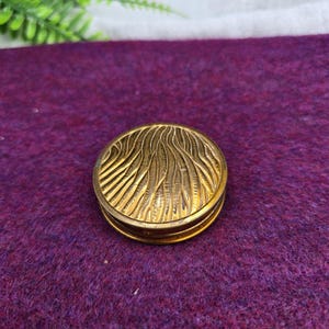 May include: A round, antique gold-colored compact with a textured, wave-like pattern on the lid. The compact is closed and appears to be made of metal. The background is a deep purple fabric.