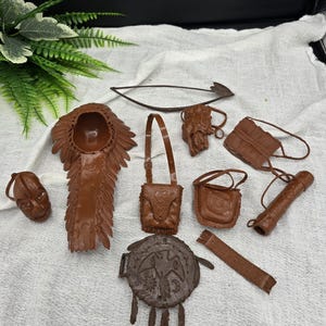 May include: A collection of brown miniature Native American-themed accessories, including a headdress, pouches, a bow and arrow, and a shield with a bird design. The items are arranged on a white cloth.