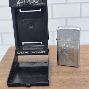 May include: A silver-tone metal Zippo lighter featuring an intricate engraved floral pattern and a smooth rectangular panel, displayed alongside an open black plastic Zippo display stand with the text Zippo U.S.A. LIGHTER and LIFETIME GUARANTEE.