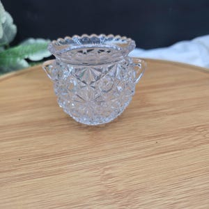 May include: A clear, small, glass sugar bowl with a diamond pattern and two small handles. The rim of the bowl has a scalloped edge. The bowl is sitting on a wooden surface.