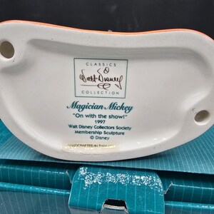 May include: A white ceramic figurine base with the text "Magician Mickey" and "On with the show!" from 1997. The Walt Disney Collectors Society membership sculpture is part of the Classics Collection. The base is orange and white.
