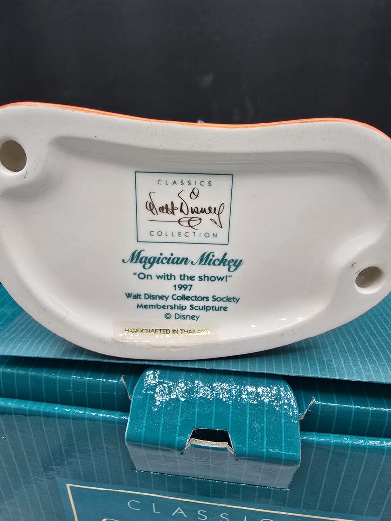 May include: A white ceramic figurine base with the text "Magician Mickey" and "On with the show!" from 1997. The Walt Disney Collectors Society membership sculpture is part of the Classics Collection. The base is orange and white.