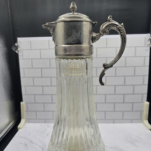 May include: A clear glass carafe with a vertical ribbed pattern and a silver-toned metal lid and handle. The metal lid features a small finial at the top, and the handle has an ornate, curved design. The glass body tapers toward the top.