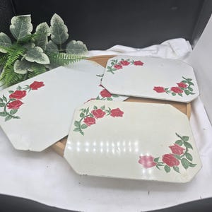 May include: Three rectangular metal trays with clipped corners, featuring an off-white base color and a decorative pattern of red roses with green leaves and stems arranged in the corners of each tray surface.