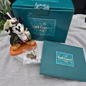 May include: A Mickey Mouse figurine in a tuxedo, top hat, and holding a cane, from the "Classics Walt Disney Collection." The figurine is on an orange base, with a matching pin and teal box.