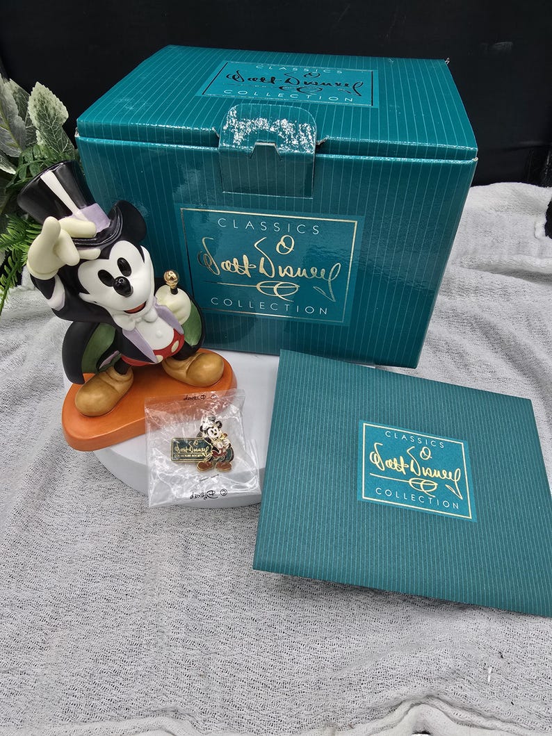 May include: A Mickey Mouse figurine in a tuxedo, top hat, and holding a cane, from the "Classics Walt Disney Collection." The figurine is on an orange base, with a matching pin and teal box.