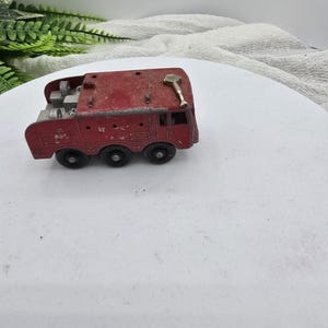 May include: A vintage red toy fire truck with six black wheels. The toy has a ladder and other details on top. The fire truck is made of metal and shows signs of wear, suggesting it is an antique or collectible item.