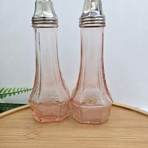 May include: A pair of vintage pink glass salt and pepper shakers with silver-colored metal lids. The shakers have a decorative floral pattern and are displayed on a wooden tray. The shakers are approximately 5 inches tall.