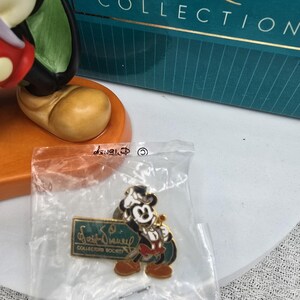 May include: A Mickey Mouse pin from the Walt Disney Collectors Society, in a clear plastic bag. The pin features Mickey in a tuxedo, holding a sword. The pin has a green and gold sign that reads "Walt Disney Collectors Society". The pin is in front of a teal box.