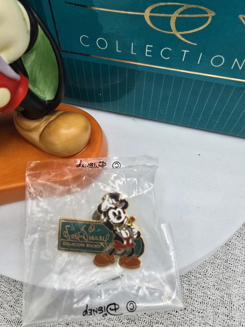 May include: A Mickey Mouse pin from the Walt Disney Collectors Society, in a clear plastic bag. The pin features Mickey in a tuxedo, holding a sword. The pin has a green and gold sign that reads "Walt Disney Collectors Society". The pin is in front of a teal box.
