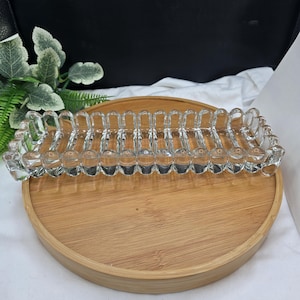 May include: A clear glass rectangular dish with a scalloped edge, resting on a round wooden tray. The dish has a series of vertical ridges. The wooden tray has a natural wood grain pattern. The background is a mix of white and black.