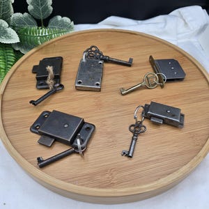 May include: A collection of four rectangular metal furniture locks and four matching keys. The locks are dark gray with mounting holes, and the keys feature various ornate bow designs. The items are arranged on a round wooden tray.