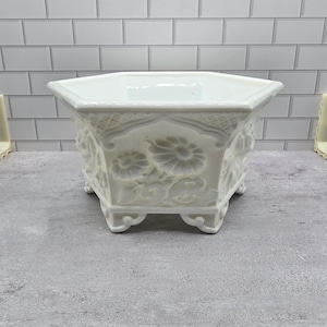 May include: A white hexagonal planter featuring embossed floral patterns on each side. The vessel has a wide, open top, a smooth interior, and is supported by six small, decorative feet at the base, providing a stable foundation for the structure.