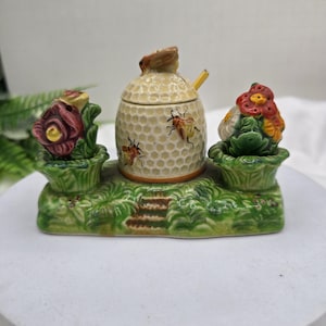 May include: A ceramic honey pot set with a beehive-shaped container, a small spoon, and two flower-shaped containers. The set is painted with yellow, green, red, and brown colors. The beehive has a honeycomb pattern and bee illustrations.
