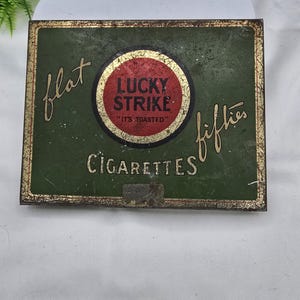 May include: Vintage Lucky Strike cigarette tin. The tin is rectangular with a green background and gold lettering. The center features a red circle with the Lucky Strike logo and the words "It's Toasted". The words "flat fifties" are on the top and bottom.