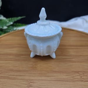 May include: A light blue, lidded trinket dish with a decorative, embossed pattern. The dish has a round body, three small feet, and a pointed finial on the lid. The dish is made of glass or ceramic.