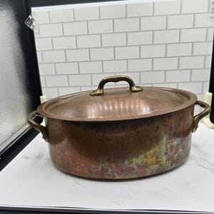 May include: An oval copper pot with a matching lid and two side handles. The metal surface displays a weathered, multi-toned patina with visible oxidation marks in shades of brown, green, and iridescent purple. The handles and lid knob are brass.