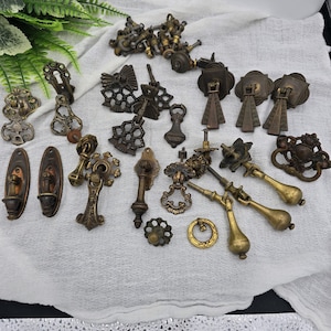 May include: A collection of antique brass and bronze drawer pulls and hardware. The assortment includes various styles, such as drop pulls, knobs, and decorative backplates. The hardware pieces are in shades of gold, bronze, and brown, with intricate designs.
