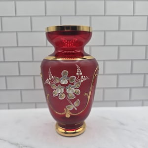 May include: A ruby red glass vase featuring a hand-painted floral motif in white and green with gold leaf accents. The vase has a flared rim and a circular base, both finished with a gold band. The body is decorated with gold filigree lines.