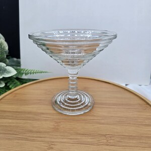 May include: A clear, stemmed glass cocktail or dessert dish. The bowl is wide and shallow, with a tiered rim. The stem is ribbed, and the base is a circular foot. The glass is sitting on a wooden tray.