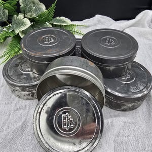 May include: A collection of vintage metal tins with lids, some open, displaying a worn, antique aesthetic. The tins are various sizes and shapes, with a circular emblem on the lids. The overall color is a muted silver and gray.