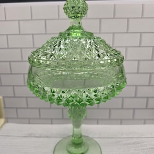 May include: A light green glass candy dish with a matching lid, featuring a diamond-cut pattern across the bowl and lid. The dish stands on a pedestal base and includes a pointed finial on top of the lid, showcasing a translucent finish.