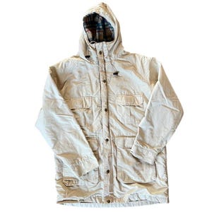 May include: A beige hooded jacket with a plaid lining visible in the hood. The jacket has multiple pockets, a button-up front, and a drawstring at the hood. The jacket is made of a lightweight material.