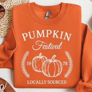 以下が含まれることがあります： オレンジ色のスウェットシャツ。前面には「PUMPKIN Festival」の文字とカボチャのイラストが描かれています。「Locally Sourced」の文字がカボチャの下にあります。スウェットシャツの横にはサングラス、靴、デニムジャケットがあります。