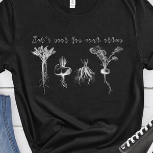 Op de afbeelding: Zwart T-shirt met een witte afbeelding van wortelgroenten en de tekst "Let's root for each other".