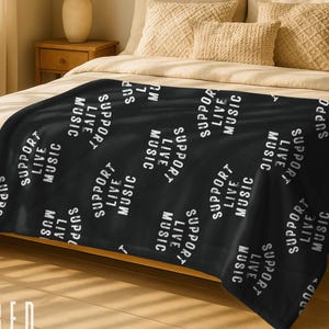 May include: Black velour blanket with the words "SUPPORT LIVE MUSIC" in white repeating across the surface. The blanket is draped over a bed with beige pillows and a wooden nightstand.
