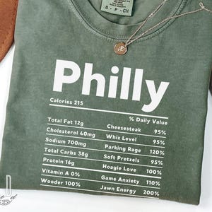 Op de afbeelding: Olijfgroen t-shirt met het woord "Philly" in grote witte letters. Het shirt heeft een voedingsfeitenlabel met voedselgerelateerde termen zoals "Cheesesteak" en "Soft Pretzels". Een gouden ketting met een hanger is over het shirt gedrapeerd.