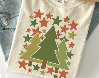 Puede incluir: Camiseta de color marfil con un gráfico de tres árboles de Navidad estilizados y estrellas en tonos verdes y naranjas. La camiseta se combina con vaqueros azul claro y un sombrero color canela. El texto "COMFORT COLORS 1717 IVORY" es visible.