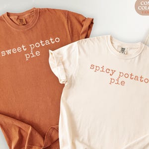 May include: Two Comfort Colors t-shirts, one in a warm brown with "sweet potato pie" printed in white, and another in ivory with "spicy potato pie" in a reddish-brown. The shirts are casual and have rolled-up sleeves.
