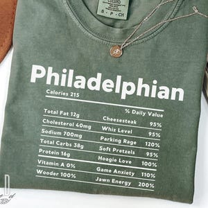 Op de afbeelding: Olijfgroen t-shirt met het woord "Philadelphian" in witte letters. Het shirt heeft een voedingswaardelabel met waarden voor cheesesteak en andere Philadelphia-thema-items. Een gouden ketting ligt over het shirt.