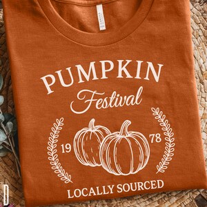 以下が含まれることがあります： オレンジ色のTシャツ。前面には「PUMPKIN Festival」の文字と2つのカボチャのイラストが描かれています。また、「1978」と「LOCALLY SOURCED」の文字も含まれています。柔らかい素材で作られています。
