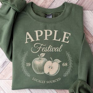 May include: Military green sweatshirt with the text "APPLE Festival" and an apple graphic. The design includes the years "19" and "68" and the words "Locally Sourced". The sweatshirt is folded on a wooden surface.