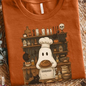 May include: Orange Bella + Canvas 3001 Autumn t-shirt featuring a ghost chef holding sourdough bread. The graphic includes a kitchen scene with shelves, candles, and a black cat. The word "Sourdough" is on the apron.
