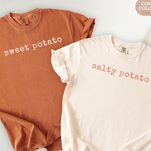 以下が含まれることがあります： 2枚のComfort Colors Tシャツ。1枚は「sweet potato」の文字が入った温かみのある茶色、もう1枚は「salty potato」の文字が入ったアイボリーです。どちらもカジュアルウェアで、柔らかいコットン製で袖がロールアップされています。