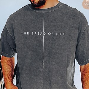 Könnte beinhalten: Dunkelgraues T-Shirt mit dem weißen Text "THE BREAD OF LIFE" über einer vertikalen weißen Linie. Der Text "JOHN 6:35" ist vertikal aufgedruckt. Das Shirt ist ein Comfort Colors 1717 Pepper.