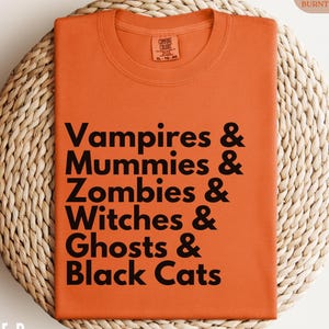 May include: Burnt orange t-shirt with the text "Vampires & Mummies & Zombies & Witches & Ghosts & Black Cats" in black. The shirt is displayed on a woven, circular surface. The shirt is a Comfort Colors 1717.
