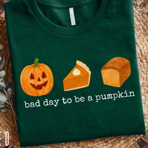 Könnte beinhalten: Dunkelgrünes T-Shirt mit einer Kürbis-, Kürbiskuchen- und Brotlaib-Grafik. Der Text "bad day to be a pumpkin" ist darunter gedruckt. Das Shirt ist ein Bella+Canvas 3001.